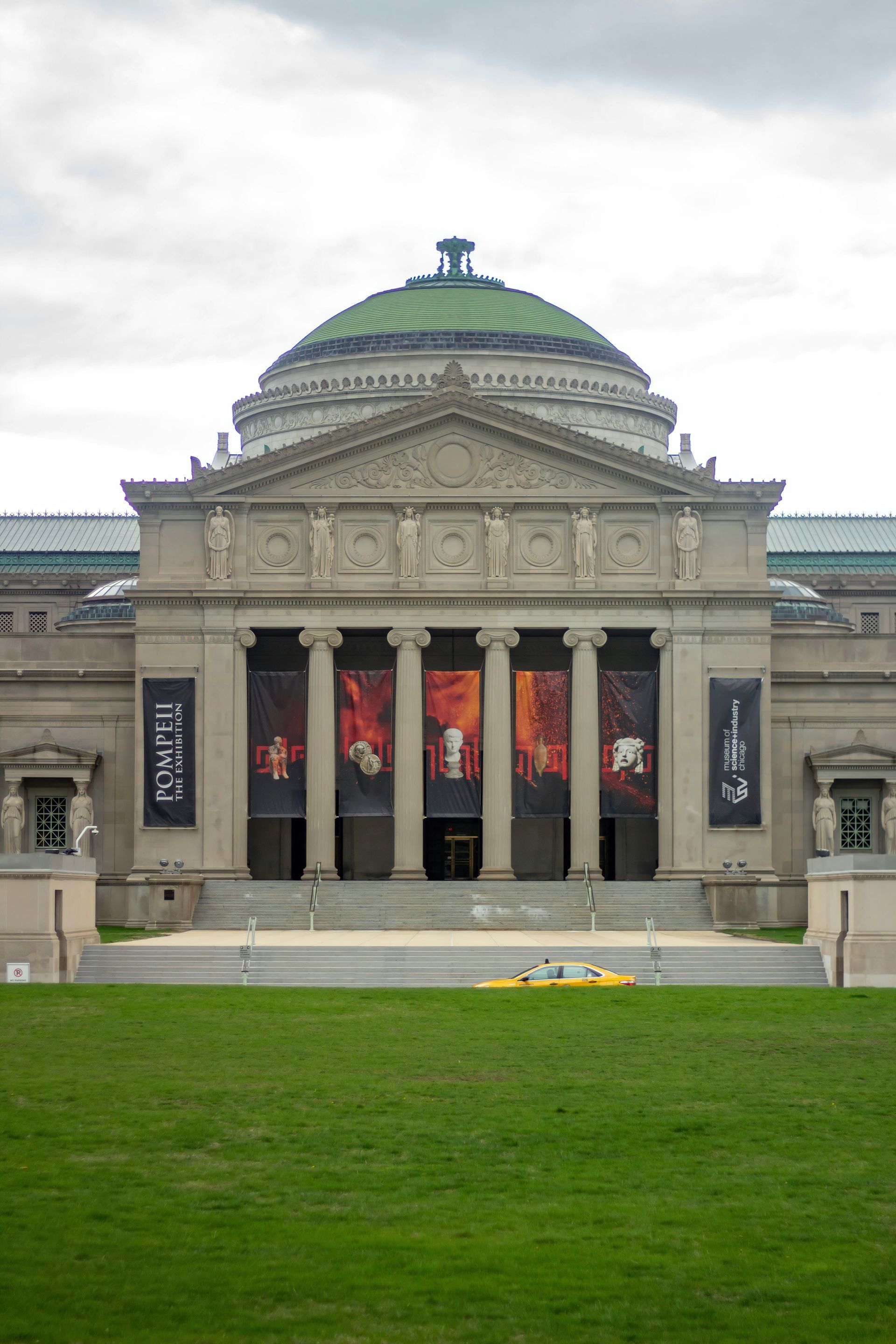 Museum with columns and a dome, banners hang above the entrance, green grass in front of the building, cloudy sky.