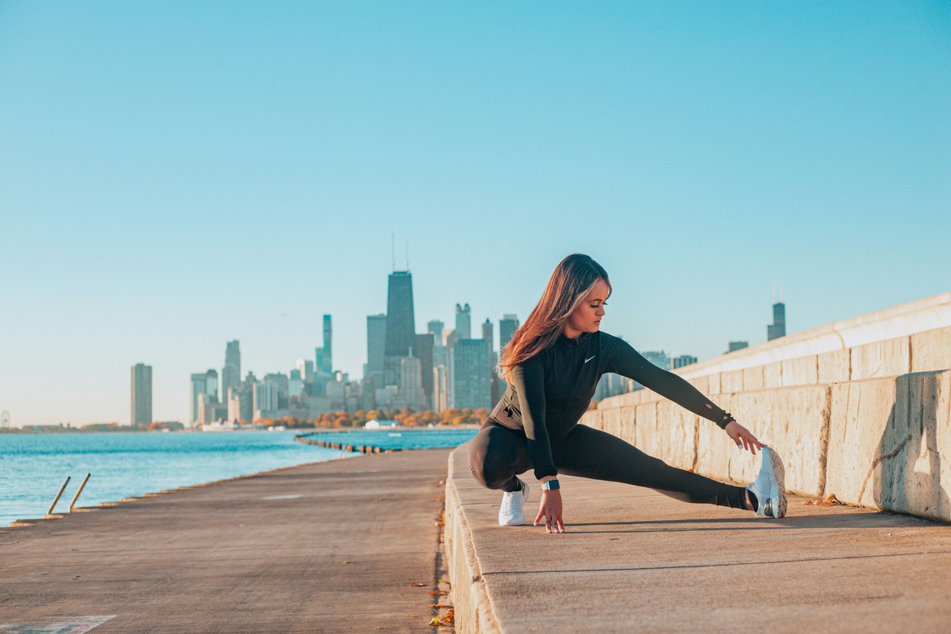 Woman stretches on a concrete wall by the water with a city skyline in the background.