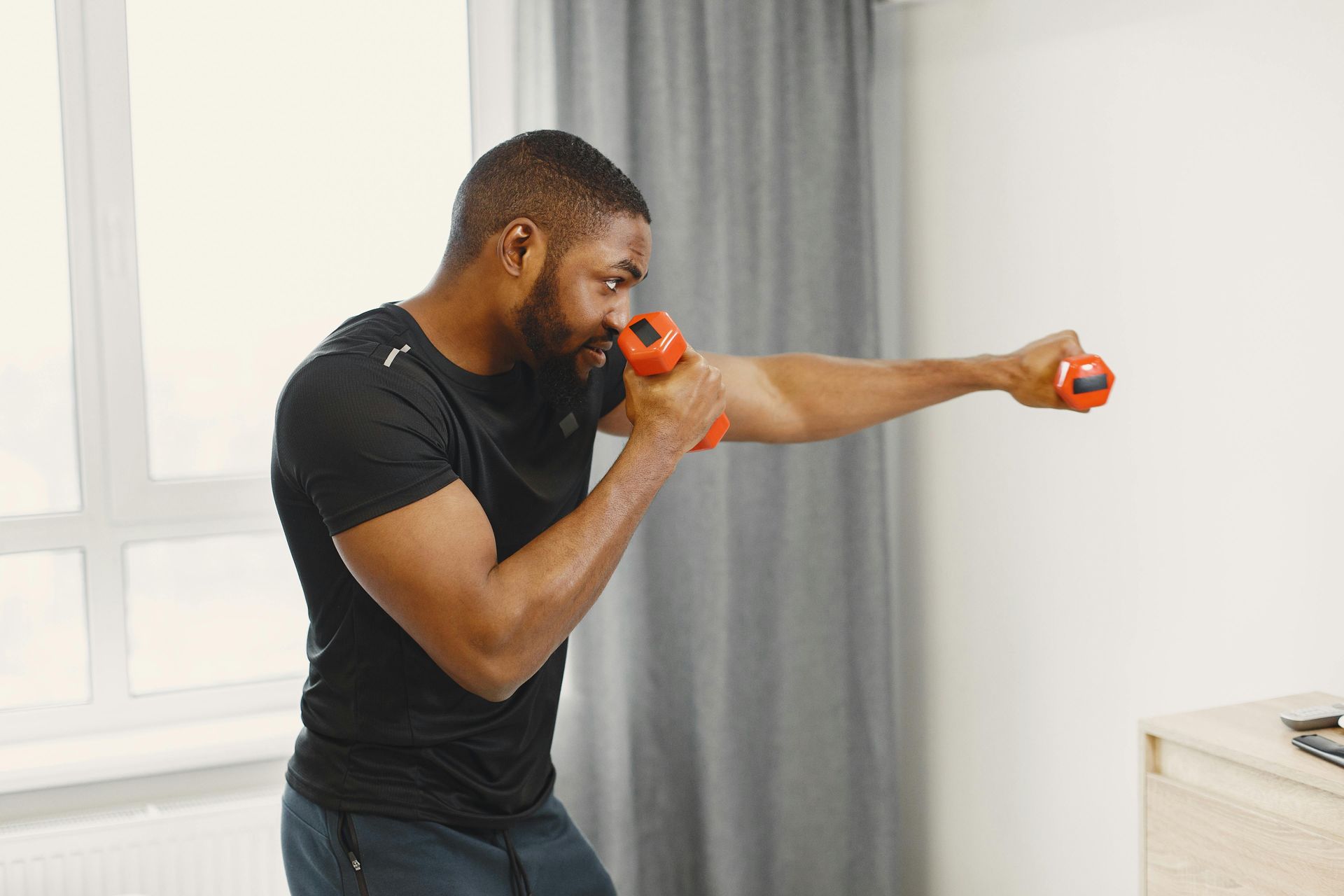A person wearing a black t-shirt performs a boxing punch exercise with small orange dumbbells in a bright room.