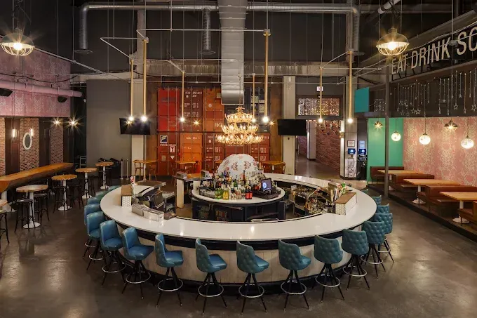 A modern, circular bar with blue stools, lit by a central chandelier in an industrial space with concrete floors.