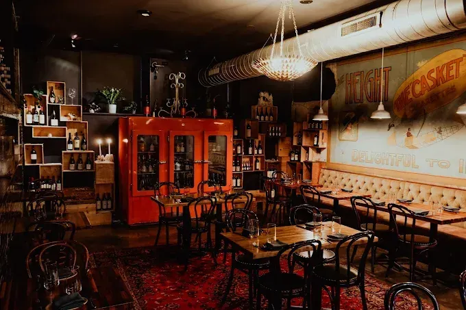 Dimly lit, cozy restaurant interior with wooden tables, black chairs, a red cabinet, and a large mural on a beige wall.
