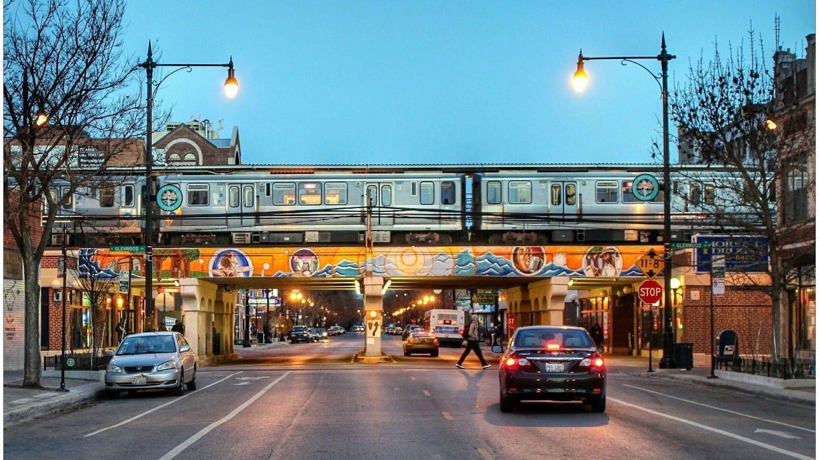 A Chicago 'L' train crosses over an urban street at dusk, with colorful murals decorating the bridge supports.