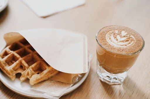 Waffle in paper wrap on a plate next to a latte with latte art, on a table.