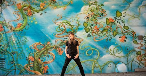 A person with long hair stands in a wide stance against a vibrant wall mural of twisting vines and orange flowers.