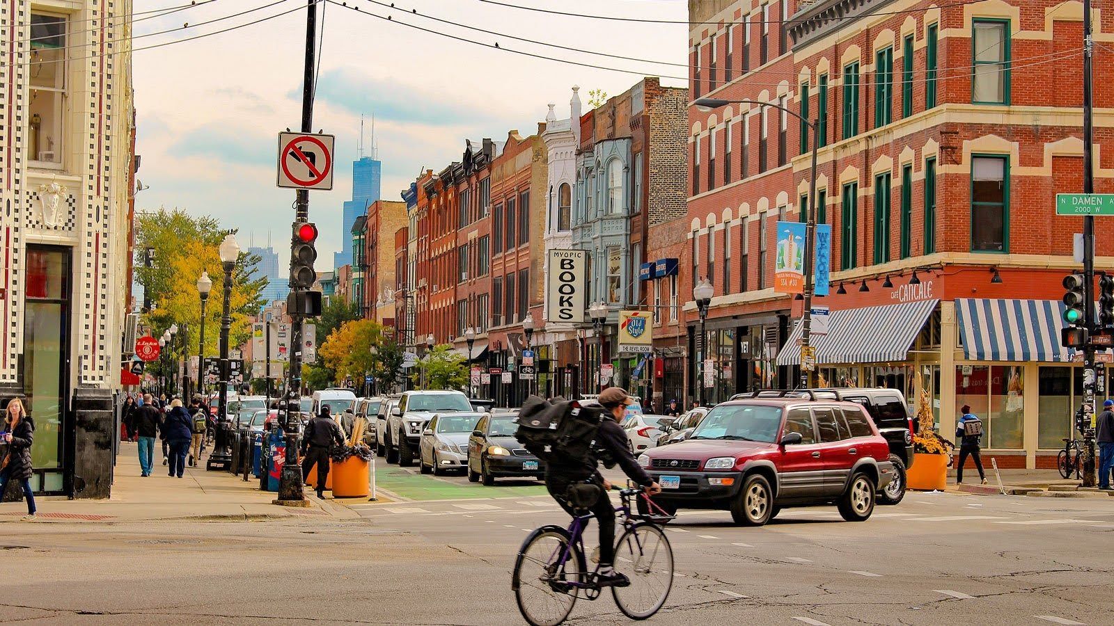 A cyclist rides across a city intersection with brick buildings, cars, and a distant skyscraper under a cloudy sky.