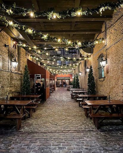 A cobblestone alleyway lit by string lights and decorated with Christmas trees, featuring tables and benches for dining.