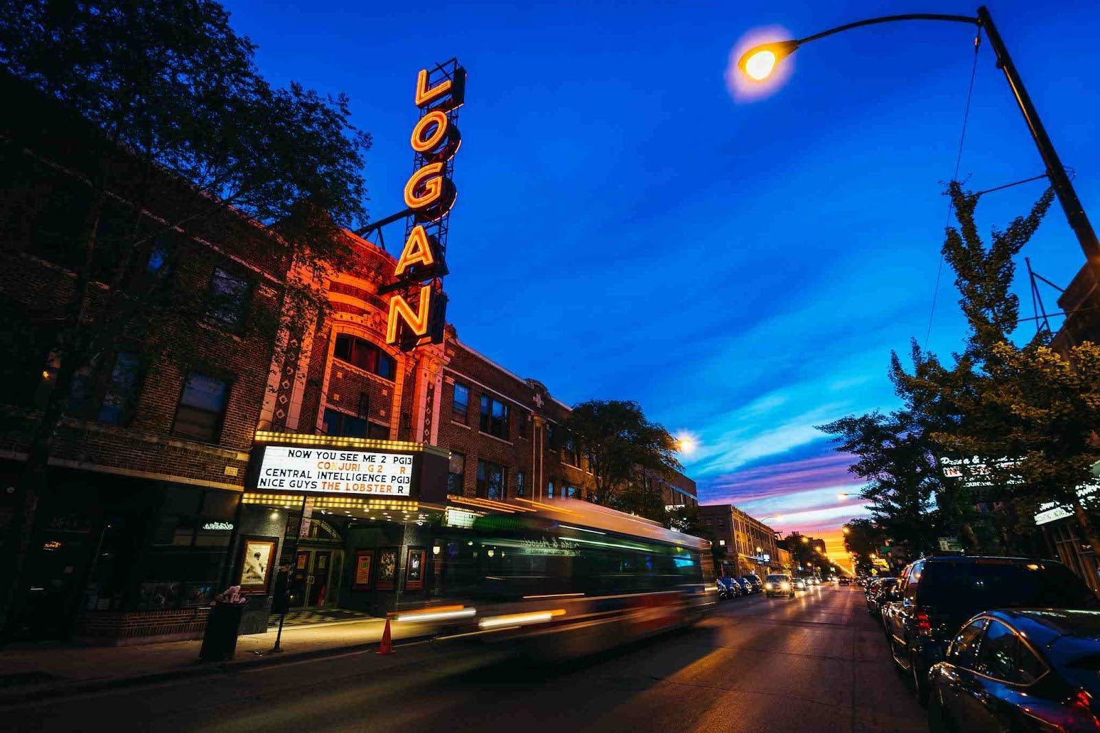 The Logan Theatre marquee lights up a Chicago street at twilight, with a blurred bus passing by and city cars parked.