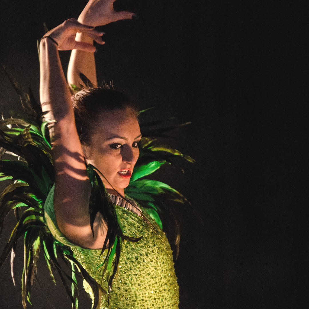 Woman in green sequined dress with feathered collar, arms raised in dance pose. Dark background.