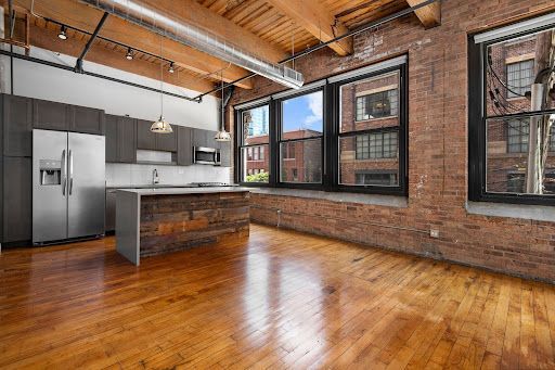 A modern open-plan kitchen and living space with brick walls, wood ceilings, hardwood floors, and large windows.