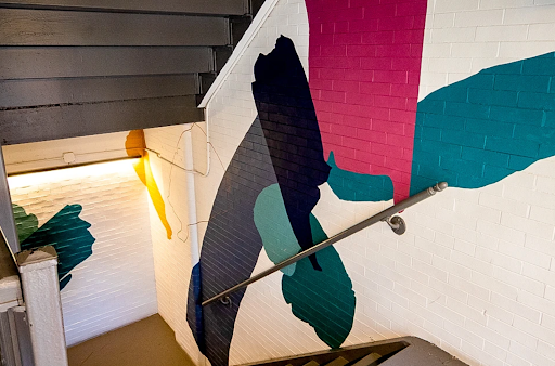 Abstract mural with magenta, teal, and dark blue shapes painted on a white brick wall by a stairwell with a metal rail.