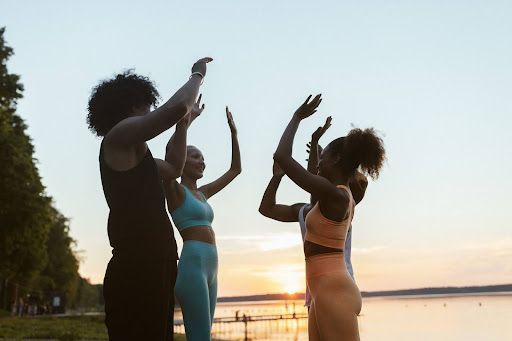 Three people in athletic wear stand in a circle by a lake at sunset, arms raised and hands touching in a high-five gesture.
