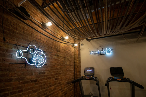 Two treadmills in a gym with exposed brick walls, industrial ceiling pipes, and glowing neon art.