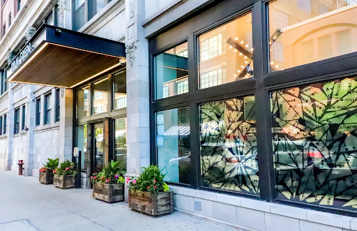 A sidewalk view of an urban building with a modern black awning, large glass windows, and potted plants lining the entry.