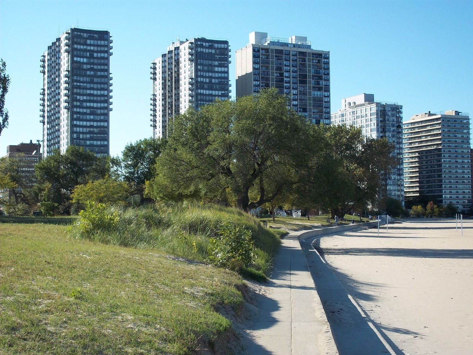 A paved path separates a grassy dune from a sandy beach, with several tall apartment towers standing in the background.