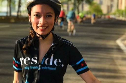 A smiling cyclist in a black and blue branded jersey and helmet, outdoors on a road with other blurred cyclists nearby.