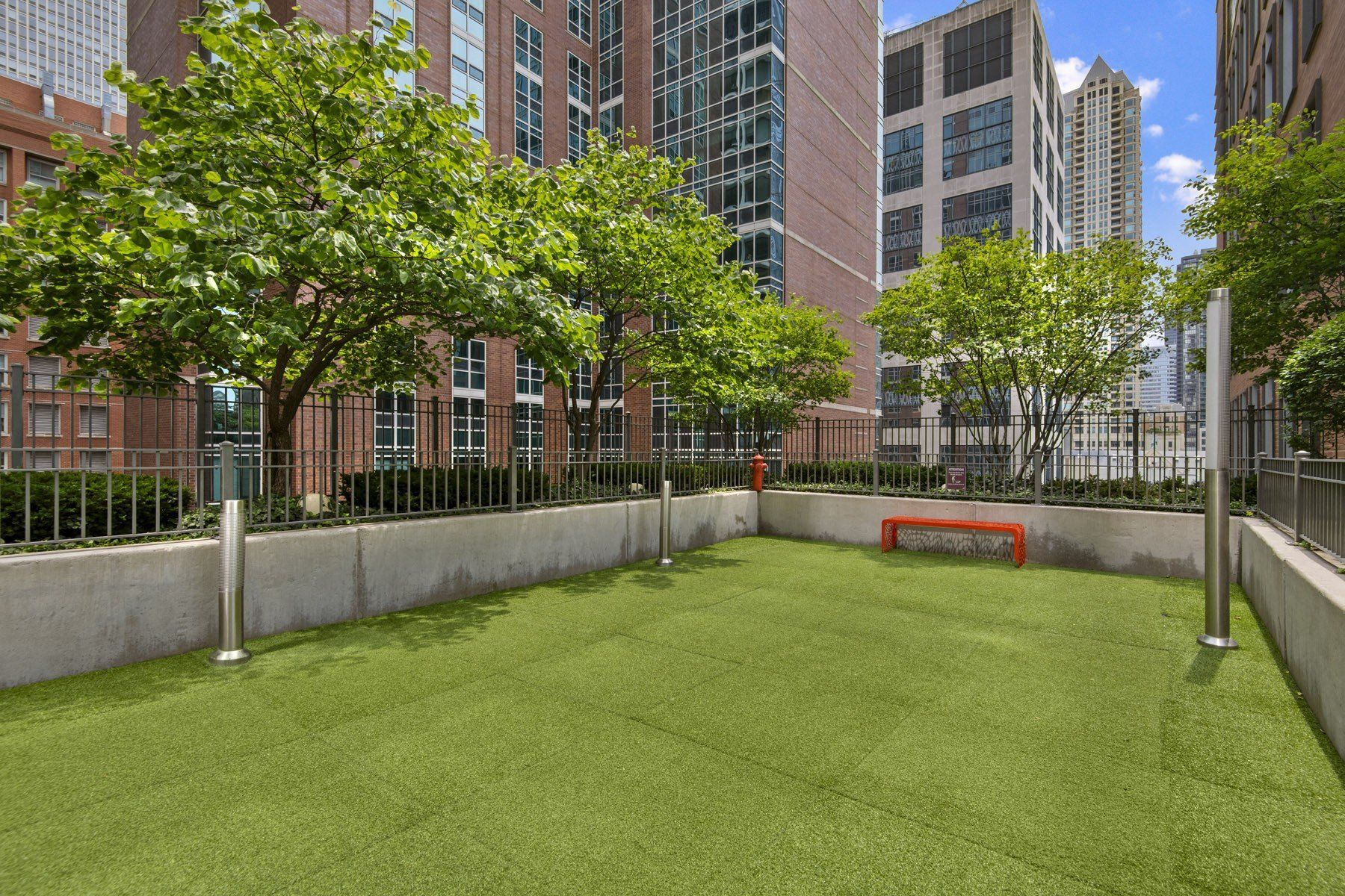 An outdoor rooftop dog run with green artificial turf, concrete walls, several trees, and a small orange bench.