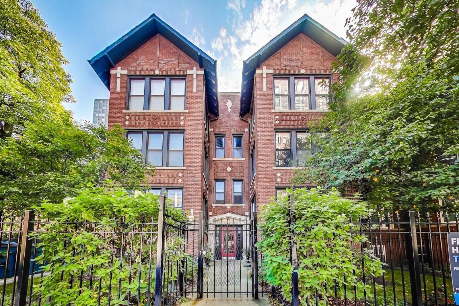 A symmetrical, three-story red brick apartment building with two gabled peaks and a central gated entrance.