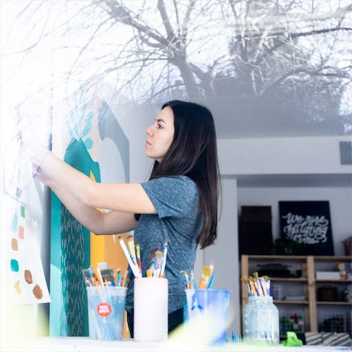 A person painting a wall mural in a studio, surrounded by paintbrushes in containers and color swatches.