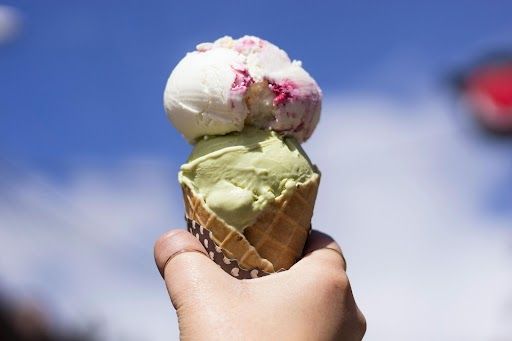Hand holding an ice cream cone with two scoops: pistachio and white with red swirls, against a blue sky.