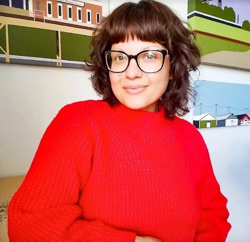 A person with shoulder-length curly hair and glasses wears a bright red sweater, smiling in front of artwork.