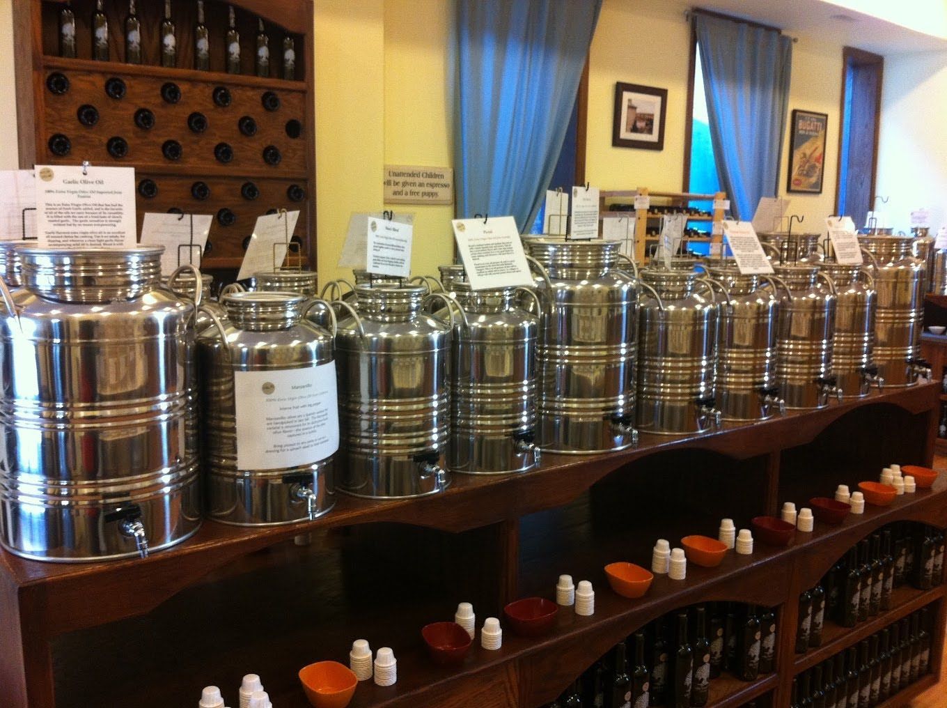 Stainless steel olive oil fusti containers lined up on a wooden store shelf with small tasting cups and sample bottles.
