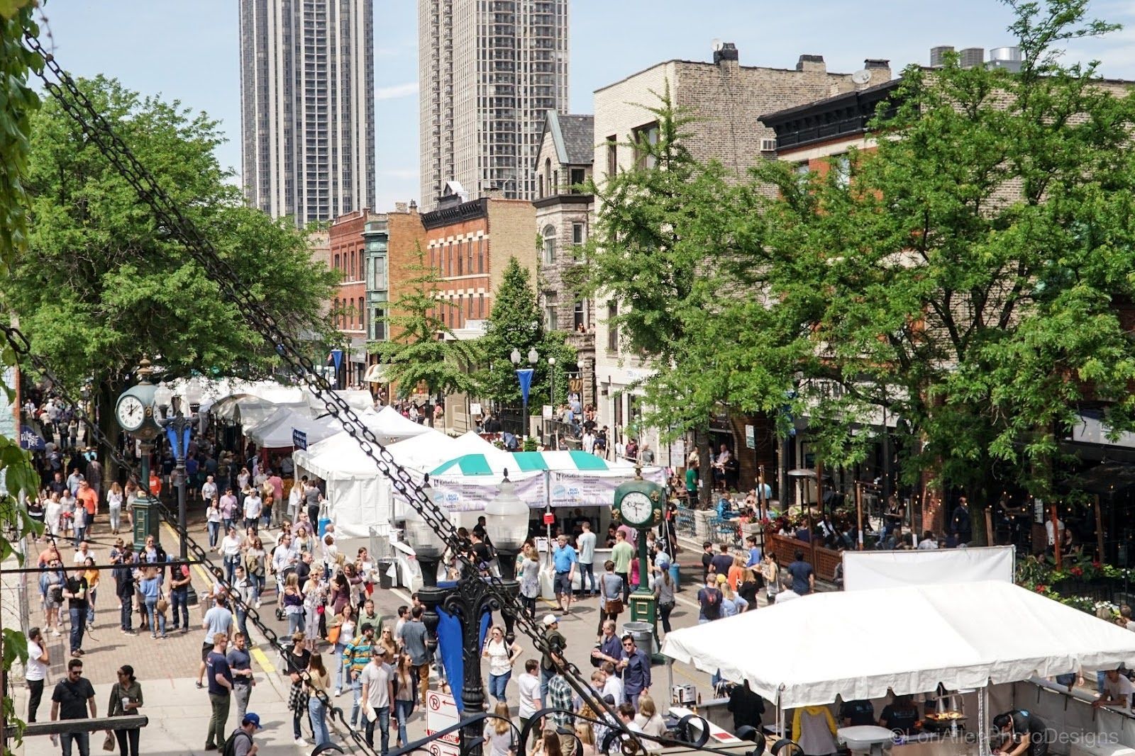 A busy street festival with many white vendor tents, crowds of people, and tall buildings under a clear blue sky.