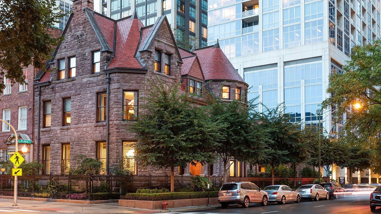 A historic, multi-story stone building with a reddish roof stands beside a modern glass high-rise on a tree-lined street.