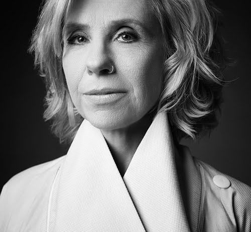 A black-and-white portrait of a person with wavy hair, wearing a white collared garment against a dark background.