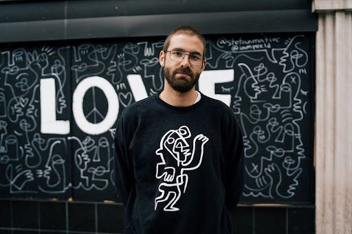 A person with a beard and glasses wears a black sweatshirt featuring line art, standing before a black mural reading 