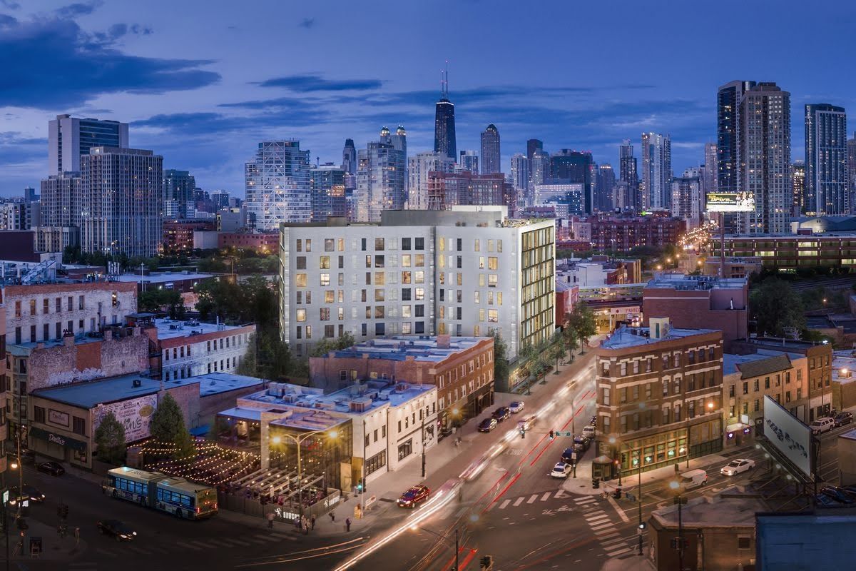 A multi-story apartment building stands above a city neighborhood at twilight with the downtown skyline in the background.