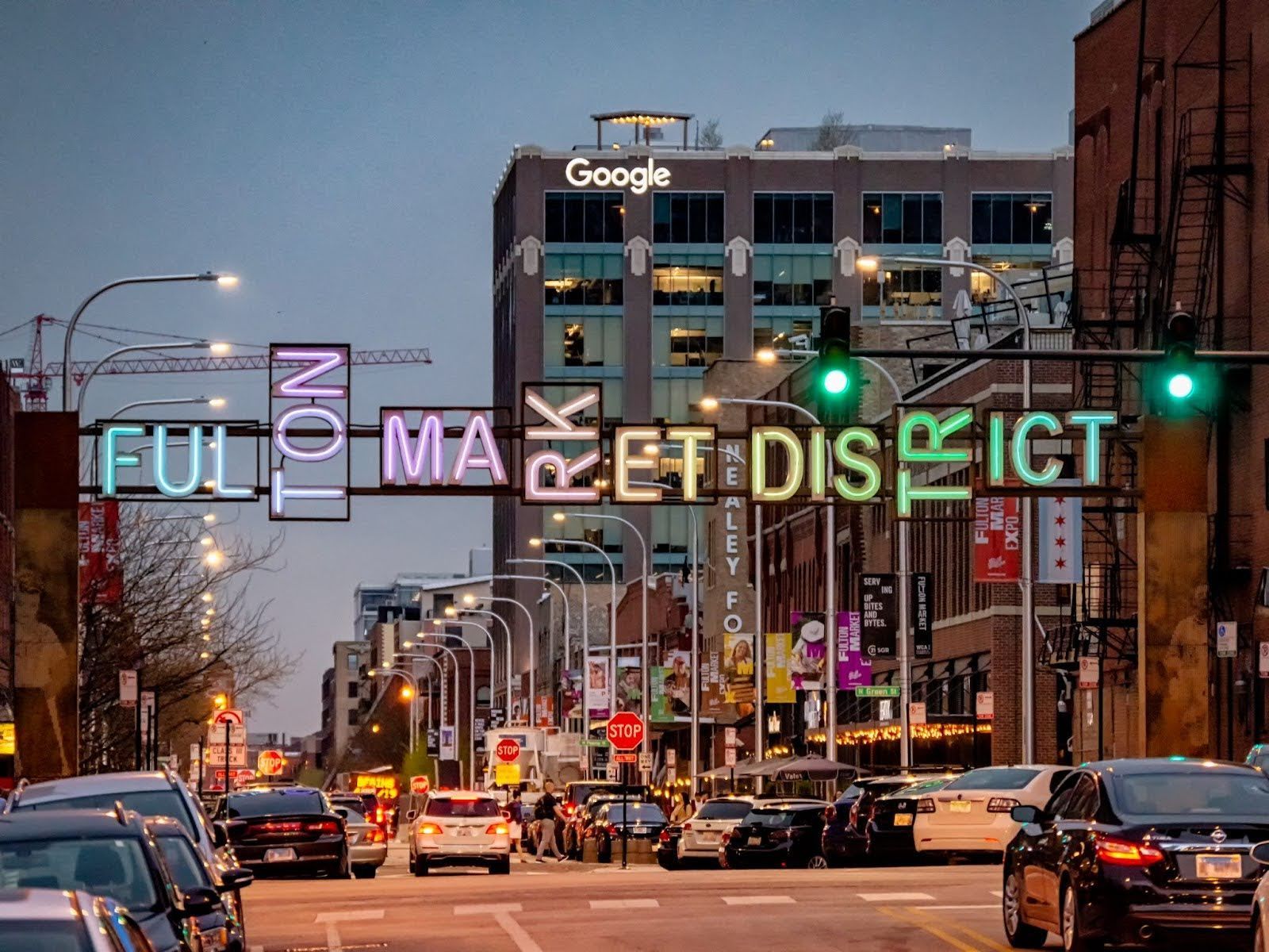 A street view of the Fulton Market District with a large, illuminated sign overhead and a Google office building beyond.