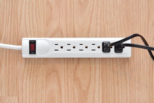 A white power strip with two black cords plugged in, lying on a light wood grain surface.