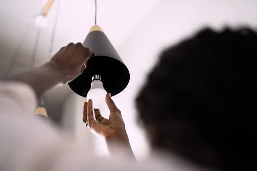 Hands reach upward to screw a white LED light bulb into a modern black pendant light fixture.