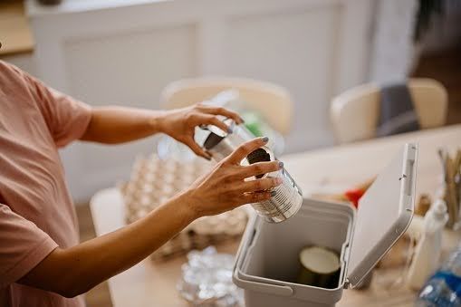 A person in a pink shirt places a metal can into a small recycling bin on a wooden table.