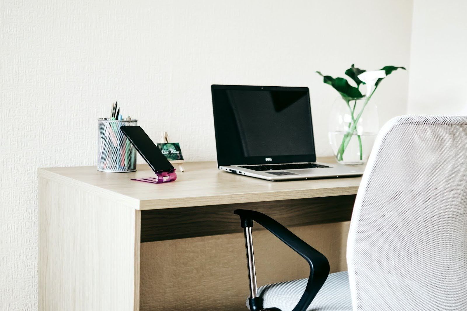 A light-colored wooden desk holds a laptop, a smartphone on a stand, a pen holder, and a small vase with flowers.