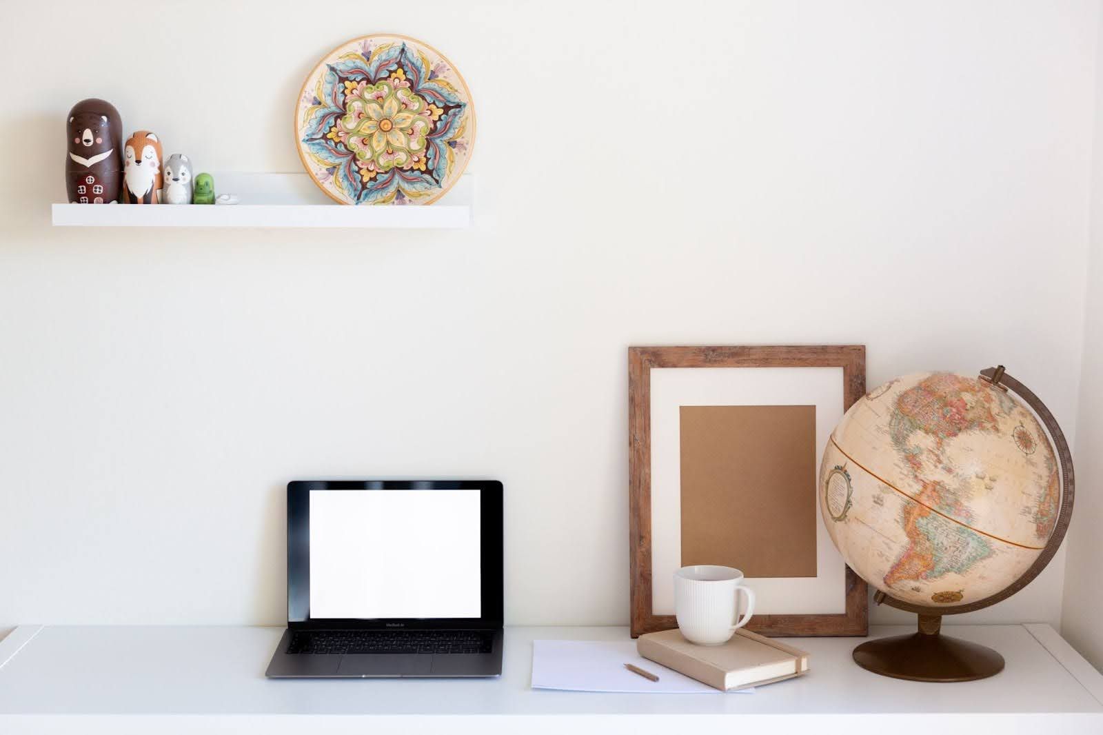 A workspace with a laptop, a globe, a picture frame, a notebook, and a small shelf with figurines on a white wall.