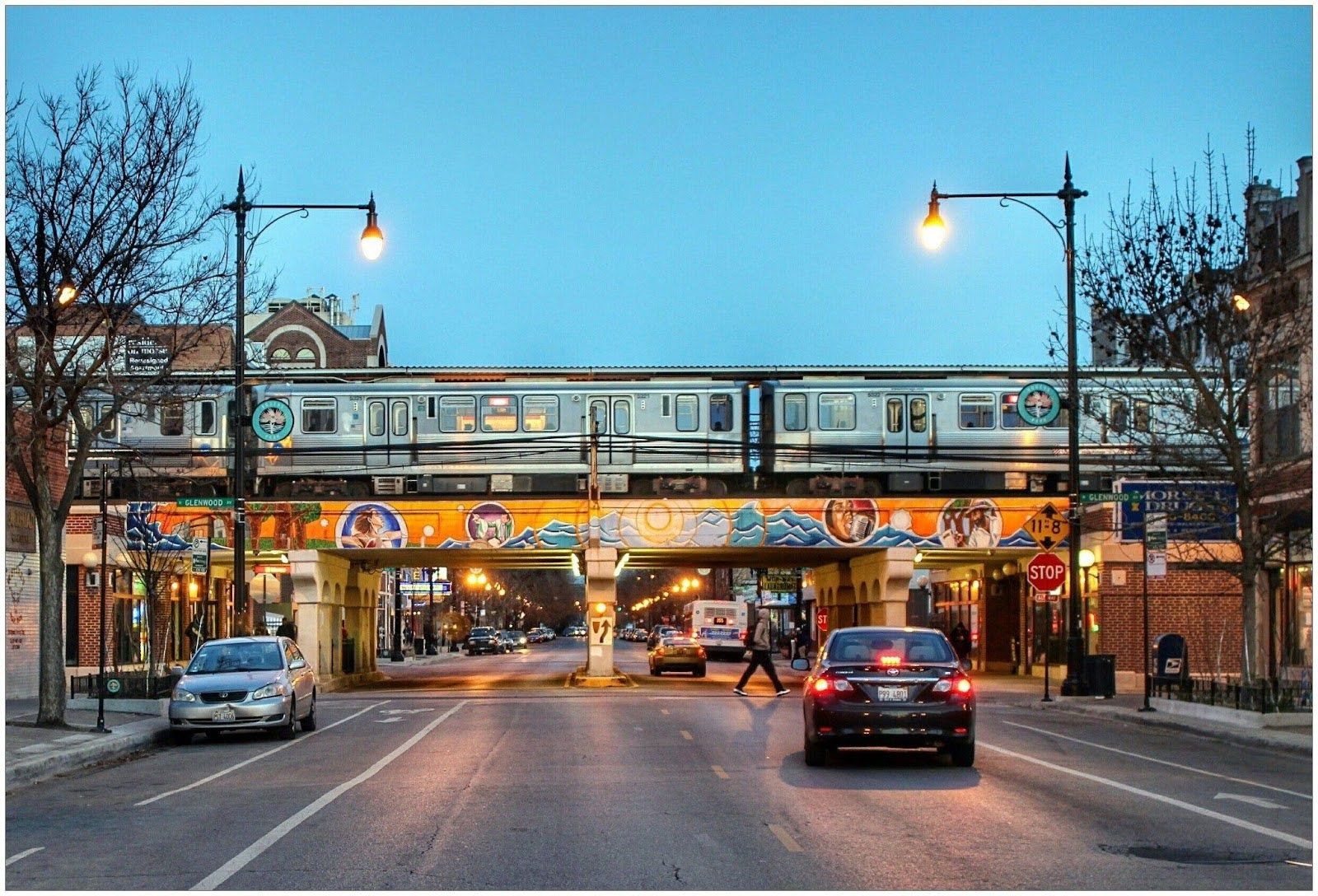 A train crosses an elevated track above a city street with cars and a pedestrian at dusk.