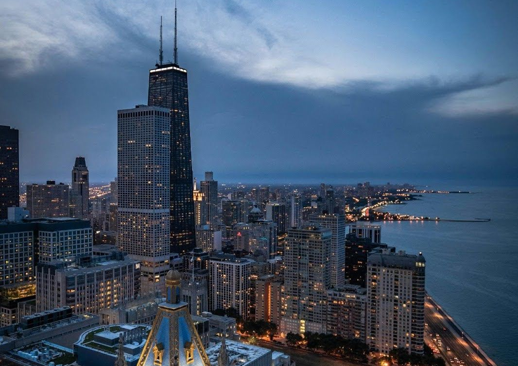 Chicago skyline at dusk featuring the John Hancock Center towering over dense city buildings and the Lake Michigan shore.