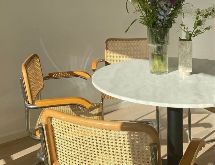 A white marble circular table with a vase of flowers sits among three wooden cane armchairs in a sunlit room.