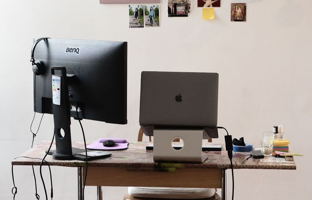 A workspace with a computer monitor, laptop on a stand, and a trash can, set against a wall with photos and notes.