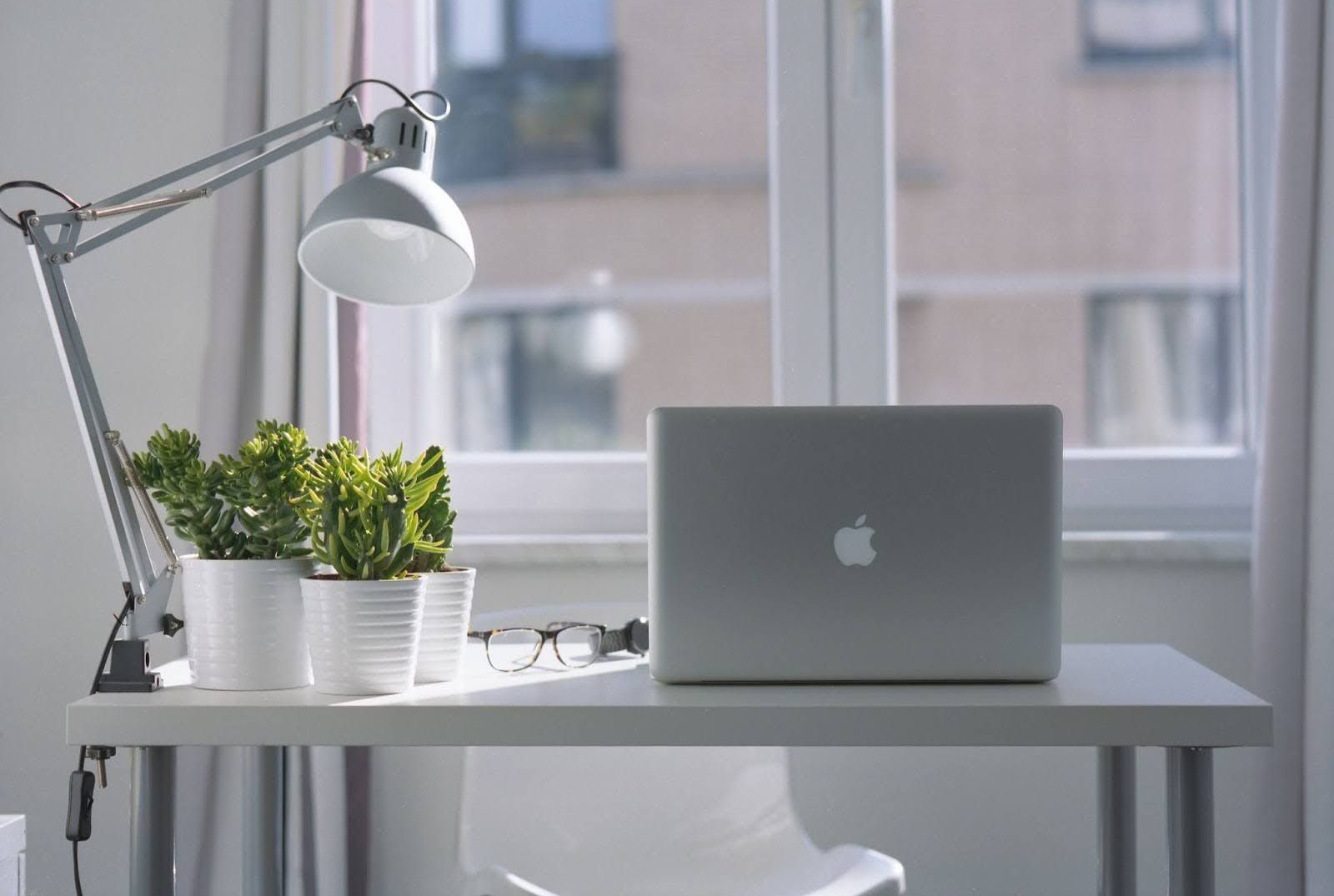 A silver laptop, a desk lamp, and small potted plants sit on a white desk in front of a bright window.