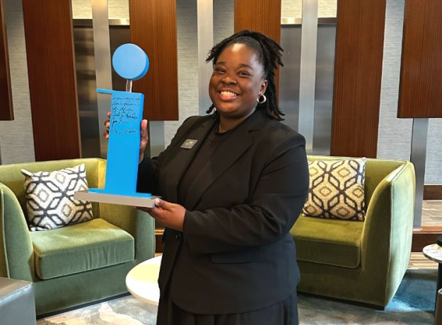 Woman holding a blue award plaque in a modern lounge with green chairs and decorative wood panels.