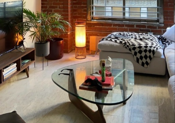 A cozy living room with exposed brick walls, a large window, a glass coffee table, and a sectional sofa with a blanket.