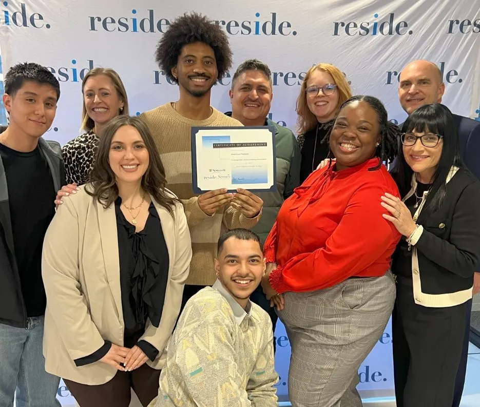 Group of people posing indoors, smiling and holding a framed certificate in front of a branded backdrop.