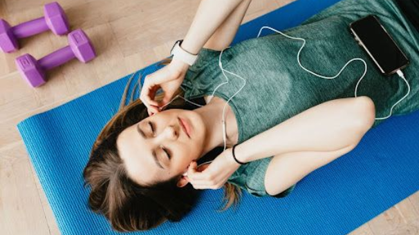 A girl listening to music, lying on top of a yoga mat with weights on the side.