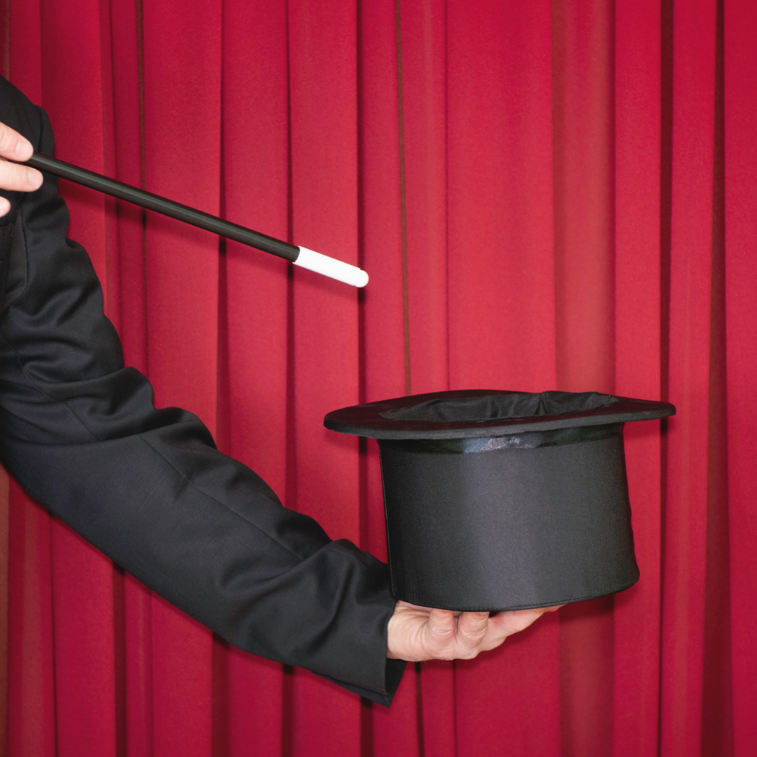 Magician in a black suit holds a top hat and wand in front of a red curtain.