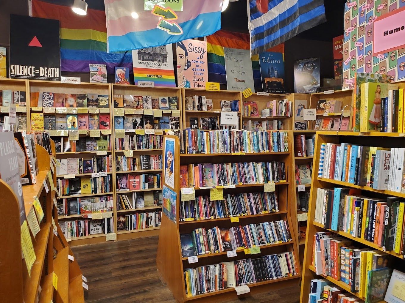 A bookstore interior with wooden shelves packed with books, featuring pride flags and posters displayed overhead.
