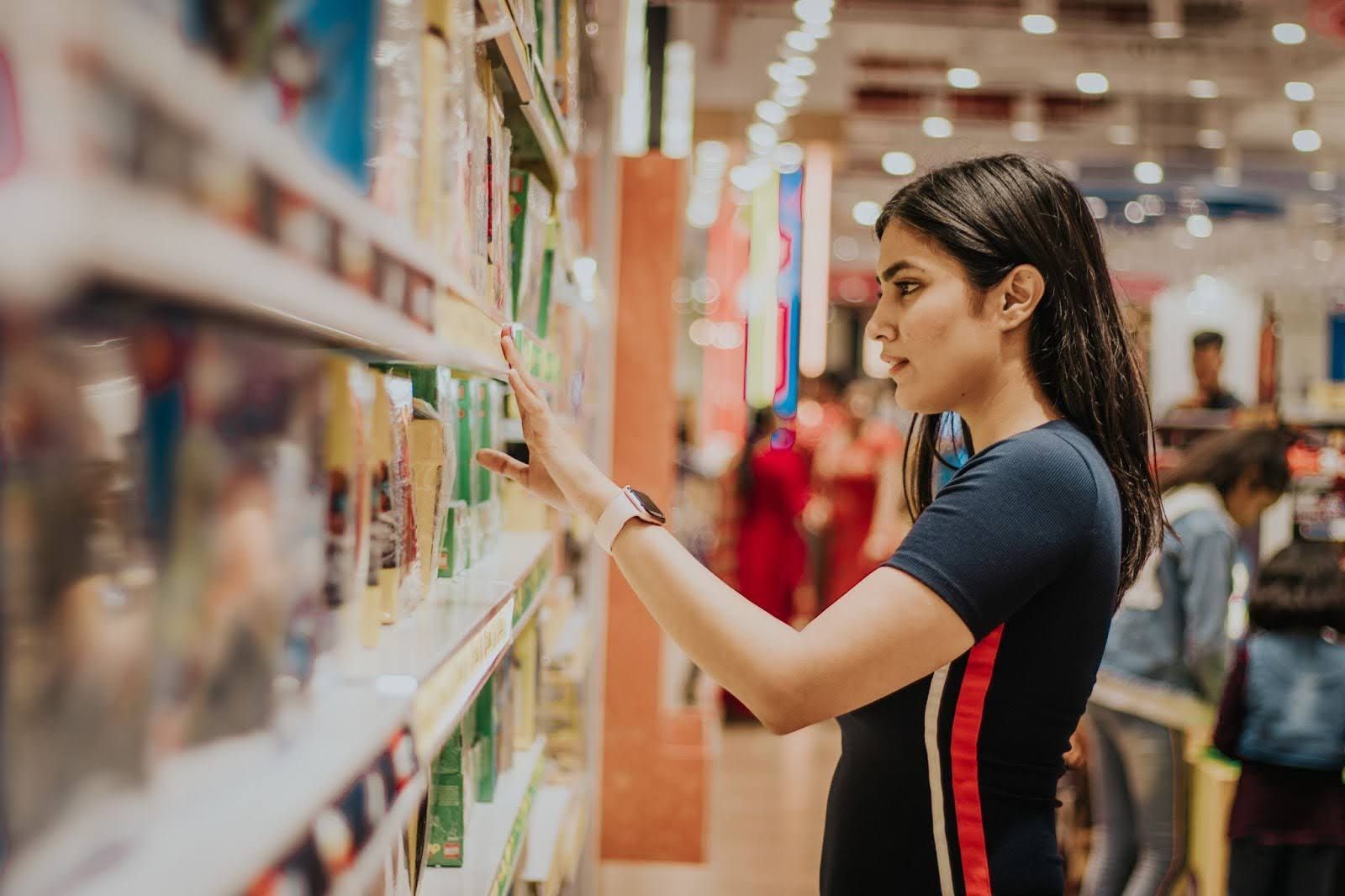 A shopper in a dark blue shirt reaches for a product on a shelf in a brightly lit, out-of-focus store aisle.