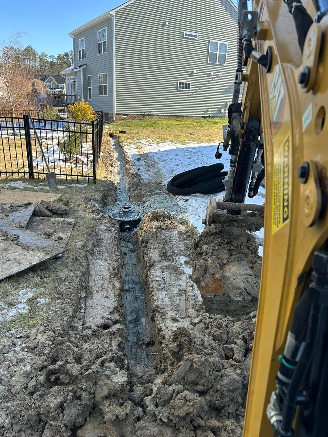 A yellow excavator digs a trench in a snowy backyard with a house in the background.