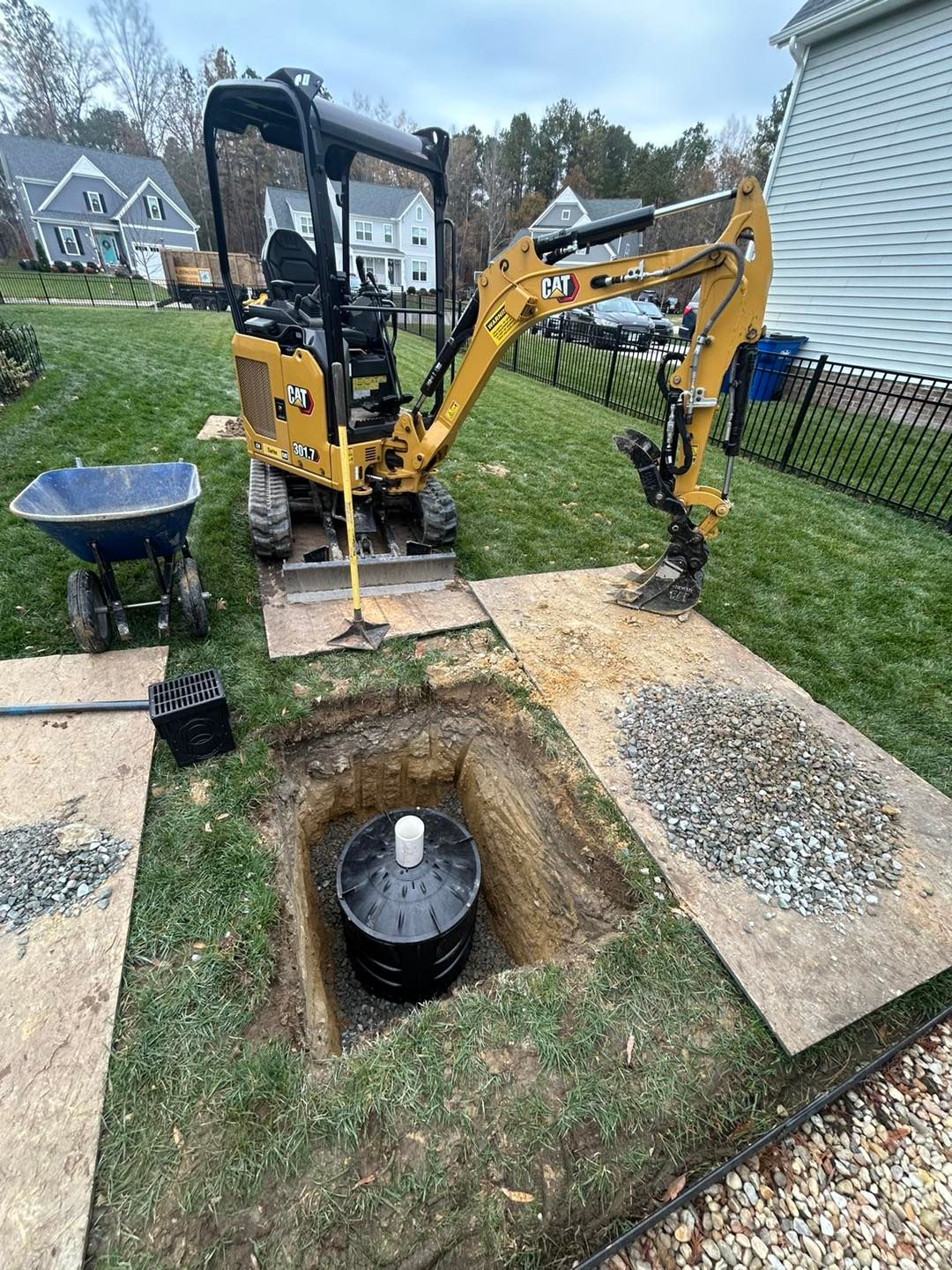 A yellow mini excavator sits in a backyard next to a hole containing a black drainage basin, with a wheelbarrow nearby.